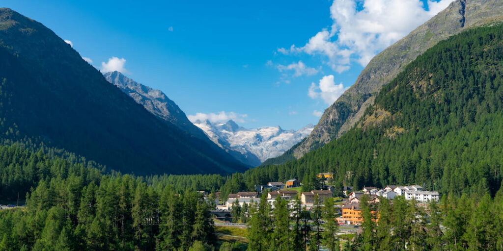 Blick auf das Dorf Pontresina im Engadin mit dem Morteratsch-Gletscher und der Berninagruppe im Hintergrund, umgeben von grünen Nadelwäldern im Sommer